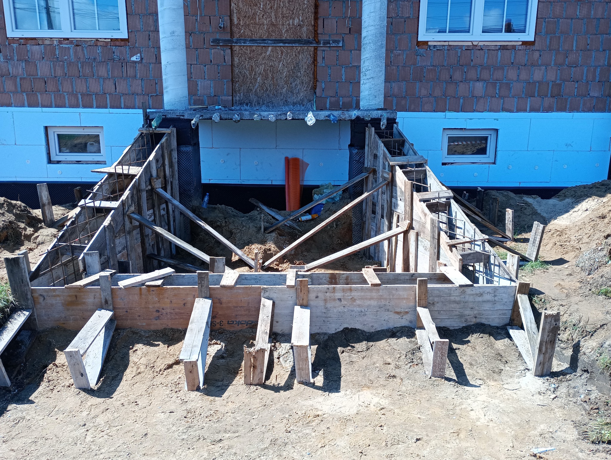 Szczegółowe ujęcie deskowania fundamentów domu z cegły, z widoczną izolacją termiczną i zbrojeniem, w trakcie budowy. Ujęcie z poziomu gruntu.