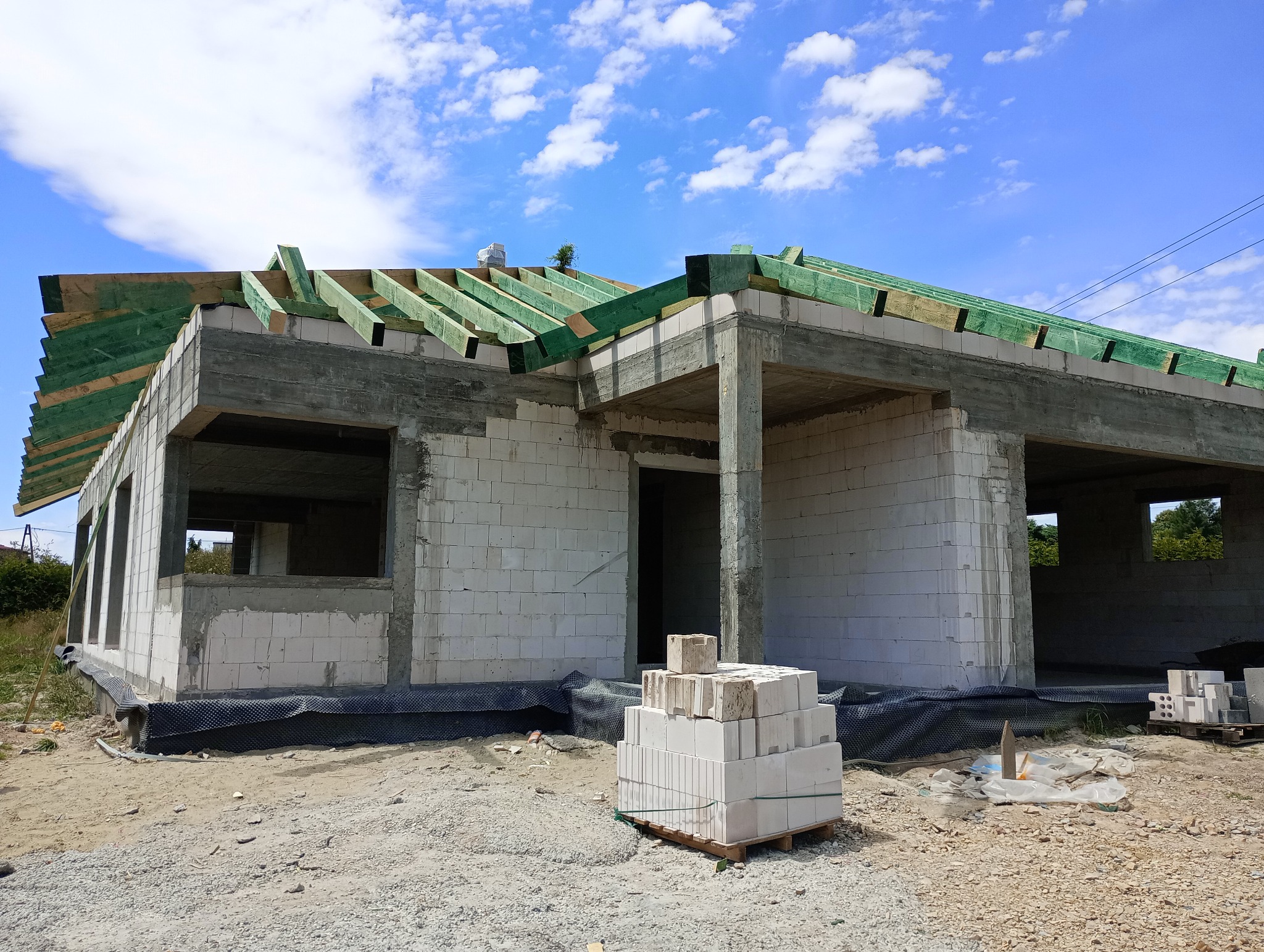 Stan surowy otwarty domu jednorodzinnego w Gogołowej. Widoczne ściany z betonu komórkowego, więźba dachowa zaimpregnowana na zielono i paleta z bloczkami.