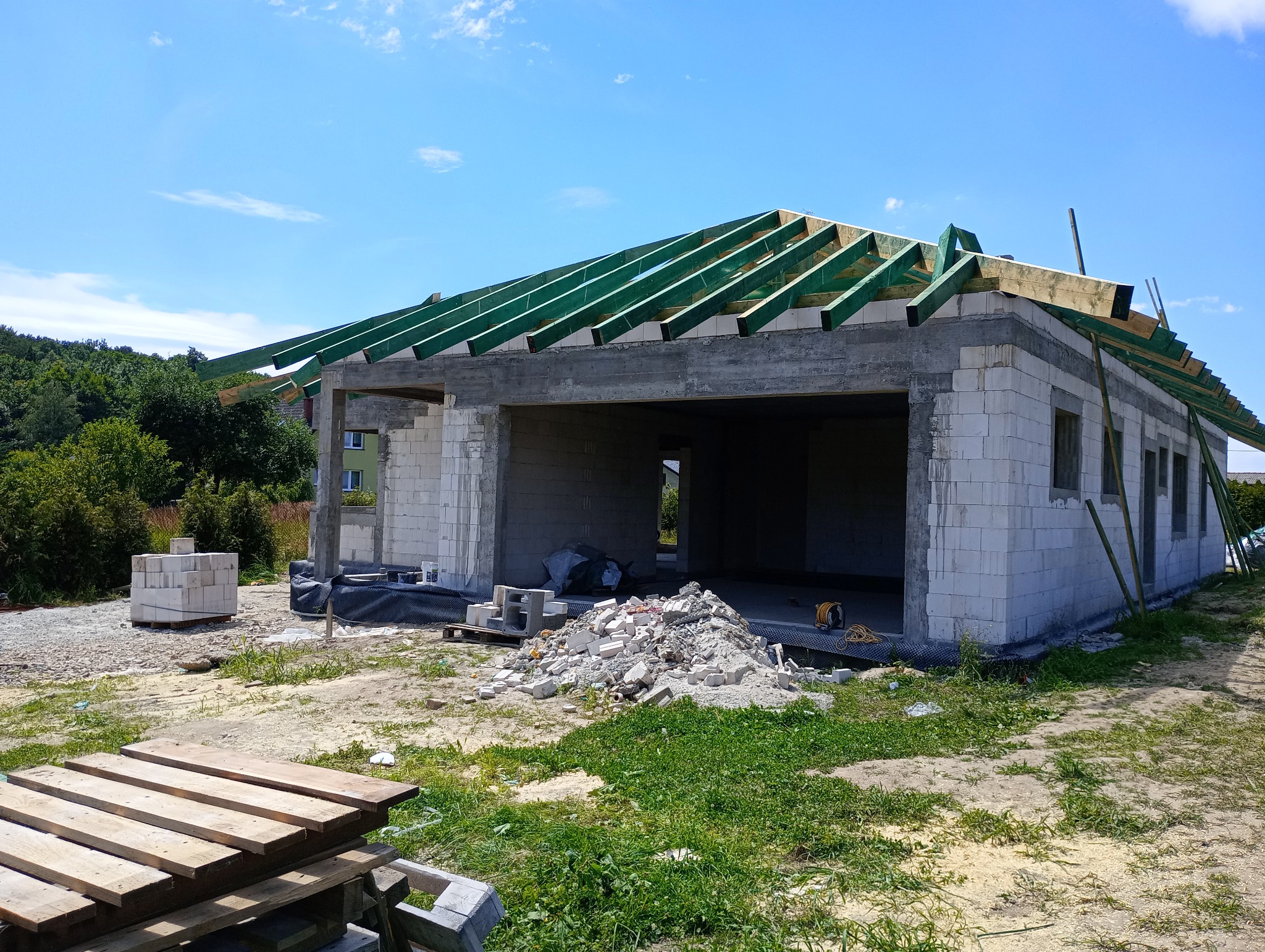 Stan surowy otwarty domu z białego bloczka, z zielonymi elementami więźby dachowej, na tle błękitnego nieba. Widoczne materiały budowlane i otoczenie w trawie.