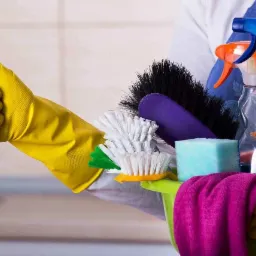 Ręka w żółtej rękawicy ochronnej pokazuje kciuk w górę, trzymając zielone wiadro z różnorodnymi akcesoriami do sprzątania: szczotki, gąbki, ściereczka i butelki ze spryskiwaczem na tle białych płytek.