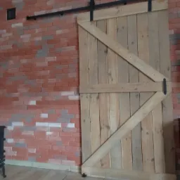 Drewniane przesuwne drzwi stodołowe z czarnym okuciem, zamontowane na tle ściany z czerwonej cegły i drewnianej podłogi.