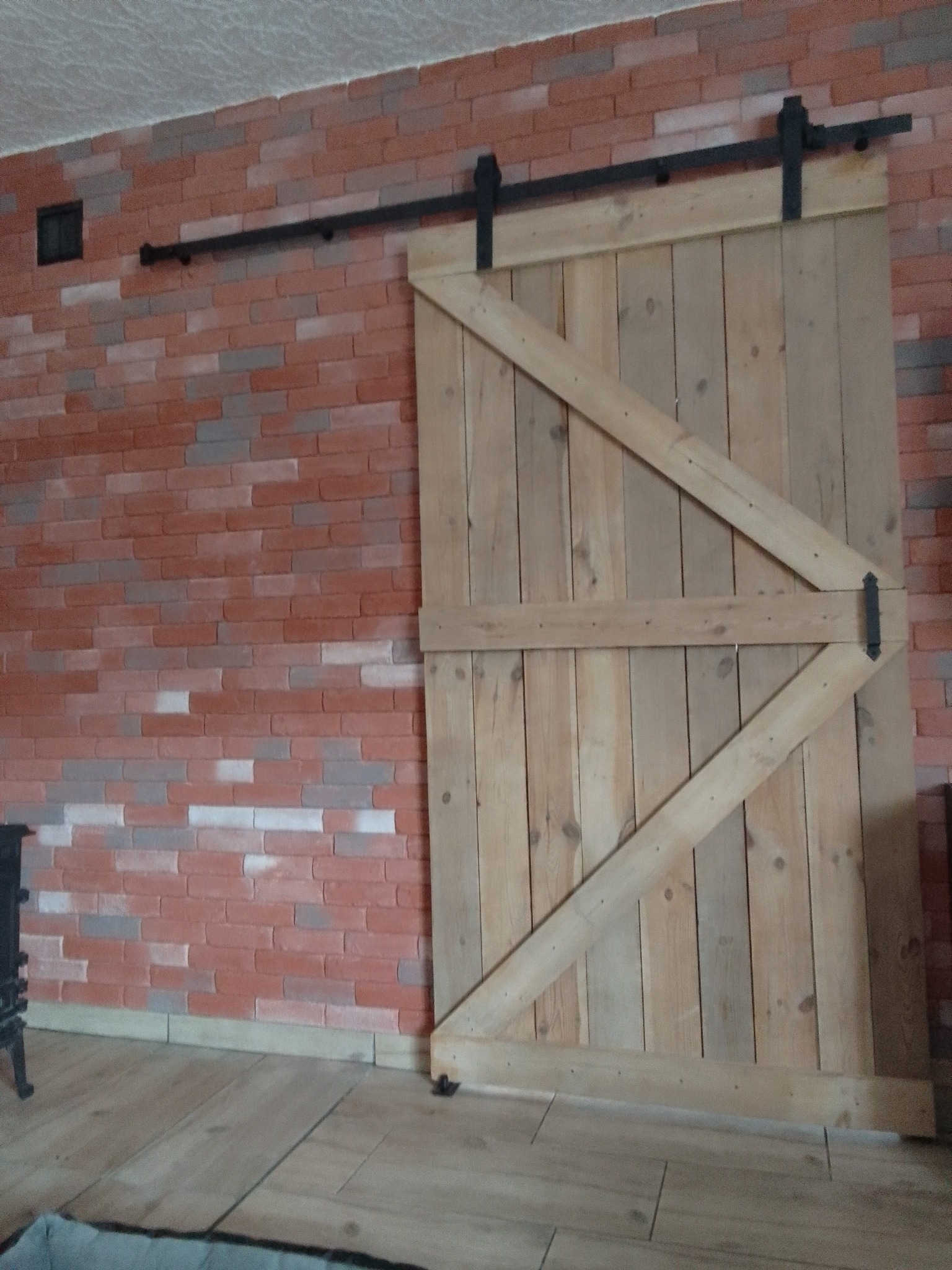 Drewniane przesuwne drzwi stodołowe z czarnym okuciem, zamontowane na tle ściany z czerwonej cegły i drewnianej podłogi.