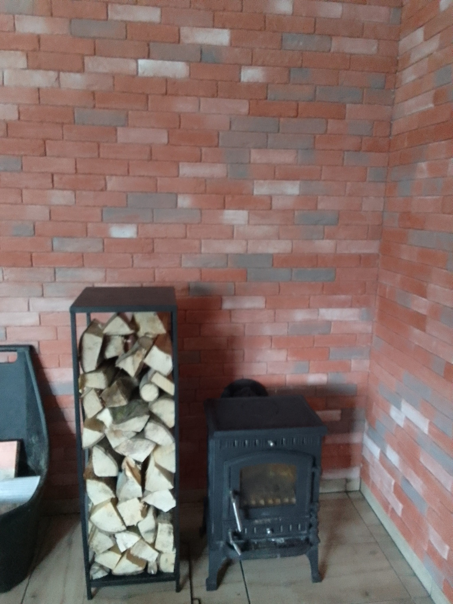 Ciemny piec wolnostojący obok metalowego stojaka wypełnionego drewnem opałowym, na tle ściany wyłożonej cegłą w odcieniach czerwieni i szarości.