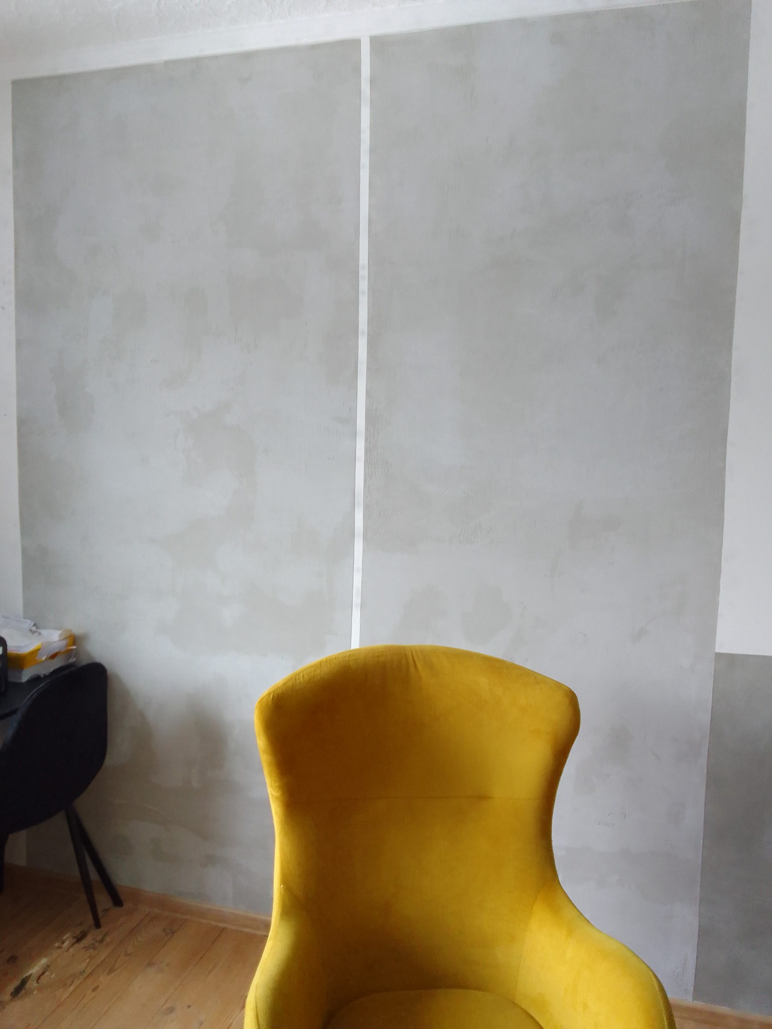 Świeżo pomalowana ściana w odcieniach szarości z pionowym paskiem taśmy malarskiej, widoczny fragment żółtego fotela welurowego i podłoga z desek.