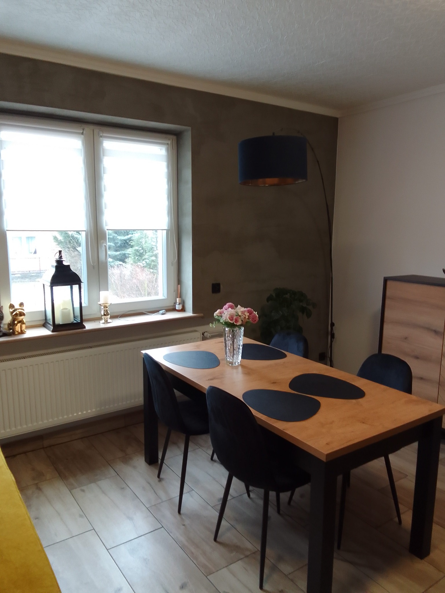 Jadalnia z drewnianym stołem z czarnymi podkładkami, aksamitnymi krzesłami, lampą łukową i dekoracjami okiennymi, ściana wykończona tynkiem dekoracyjnym.