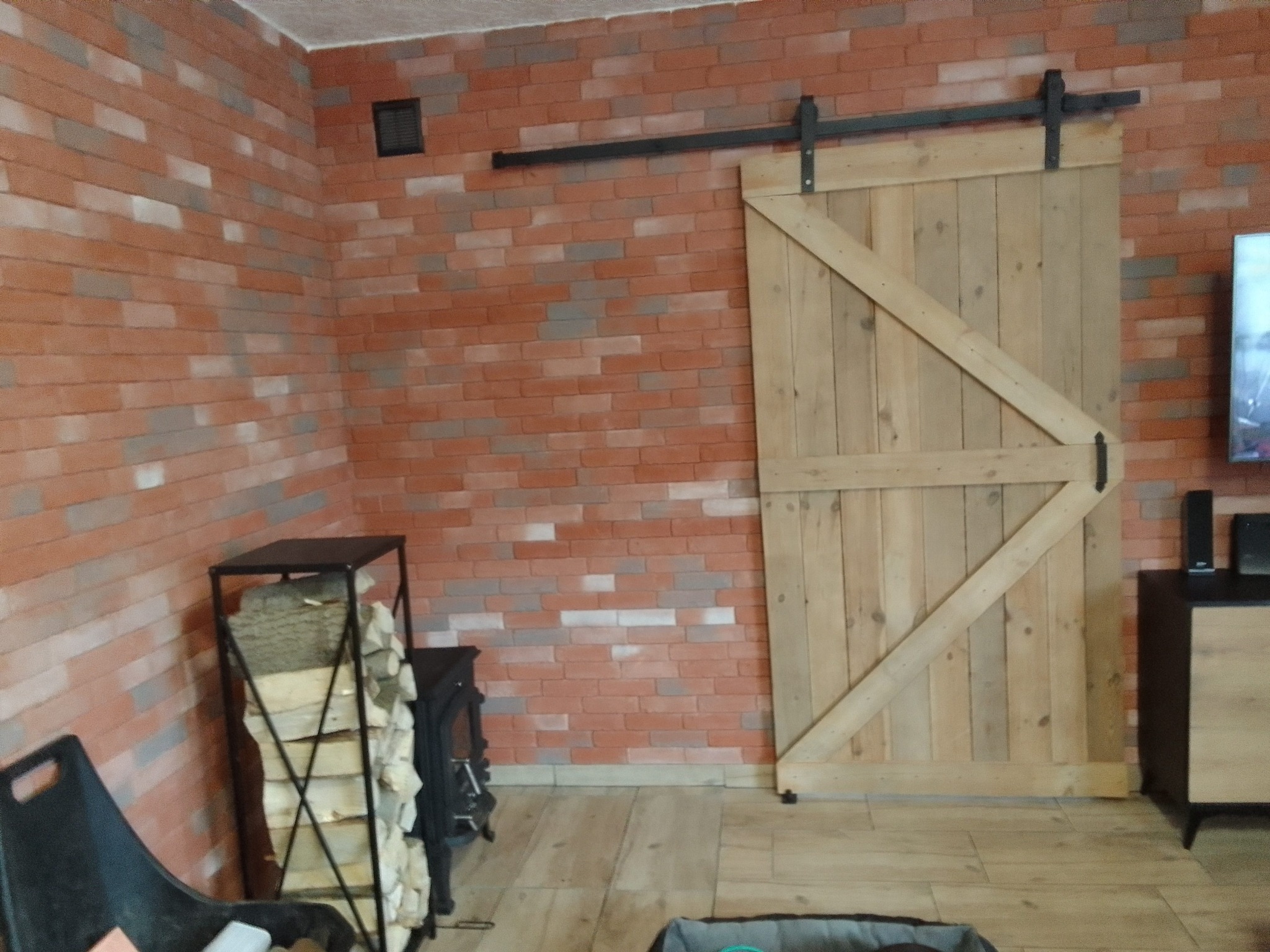 Drewniane drzwi przesuwne w stylu rustykalnym na tle ściany z cegły dekoracyjnej, widoczny czarny system przesuwny, obok kominek z zapasem drewna.