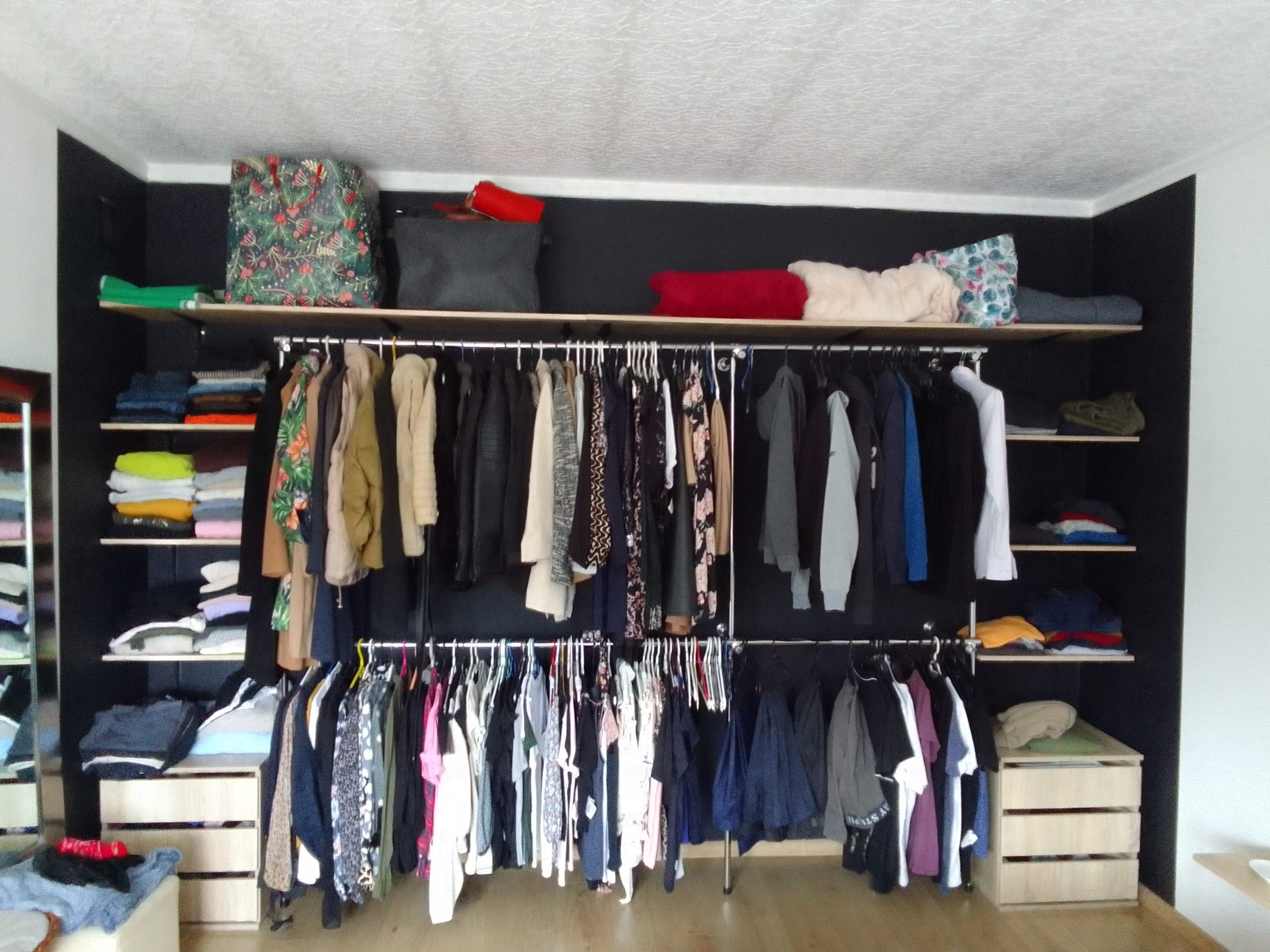 Wnętrze garderoby z czarnymi ścianami, drewnianymi półkami i dwoma rzędami ubrań na wieszakach, uzupełnione komodami z jasnego drewna.