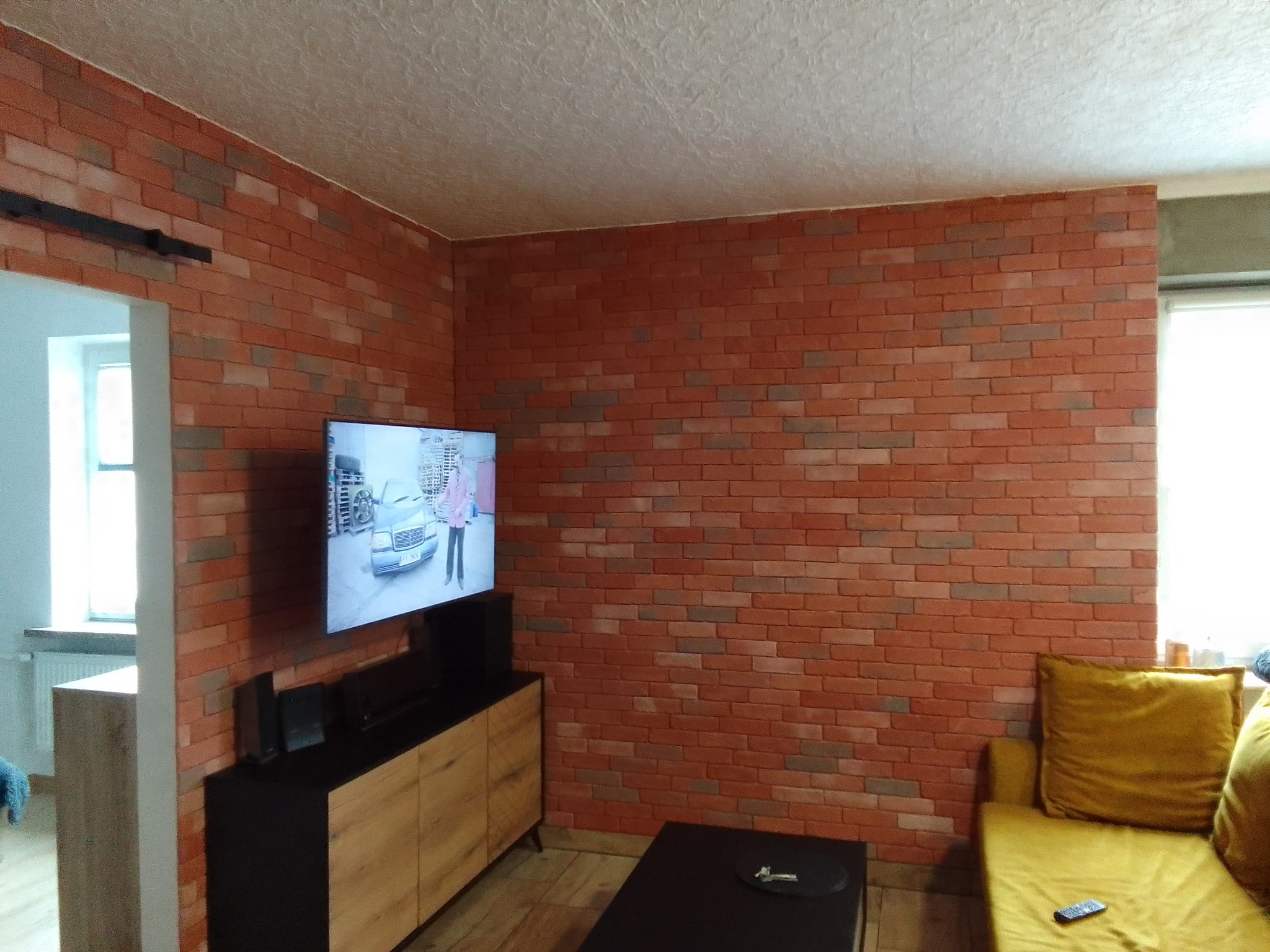Ściana wyłożona dekoracyjną czerwoną cegłą w salonie, widoczny fragment sufitu z fakturą, telewizor na szafce RTV i żółta sofa.