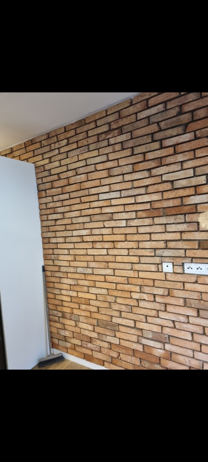 Ściana wyłożona dekoracyjną cegłą w odcieniach brązu i beżu, widoczne gniazdka elektryczne, fragment białej ściany i mop przy podłodze.