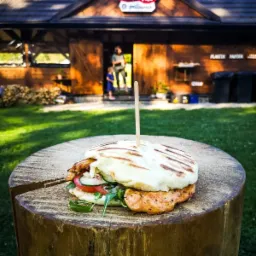 Grillowana kanapka z kurczakiem, warzywami i sosem, spięta wykałaczką, umieszczona na drewnianym pniu, w tle budynek z napisem 'grillaurus' i osoby w wejściu.