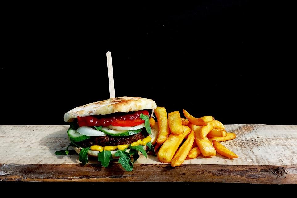 Burger z wołowiną, rukolą, ogórkiem, cebulą i pomidorem, polany sosem, podany z frytkami na drewnianej desce i czarnym tle.