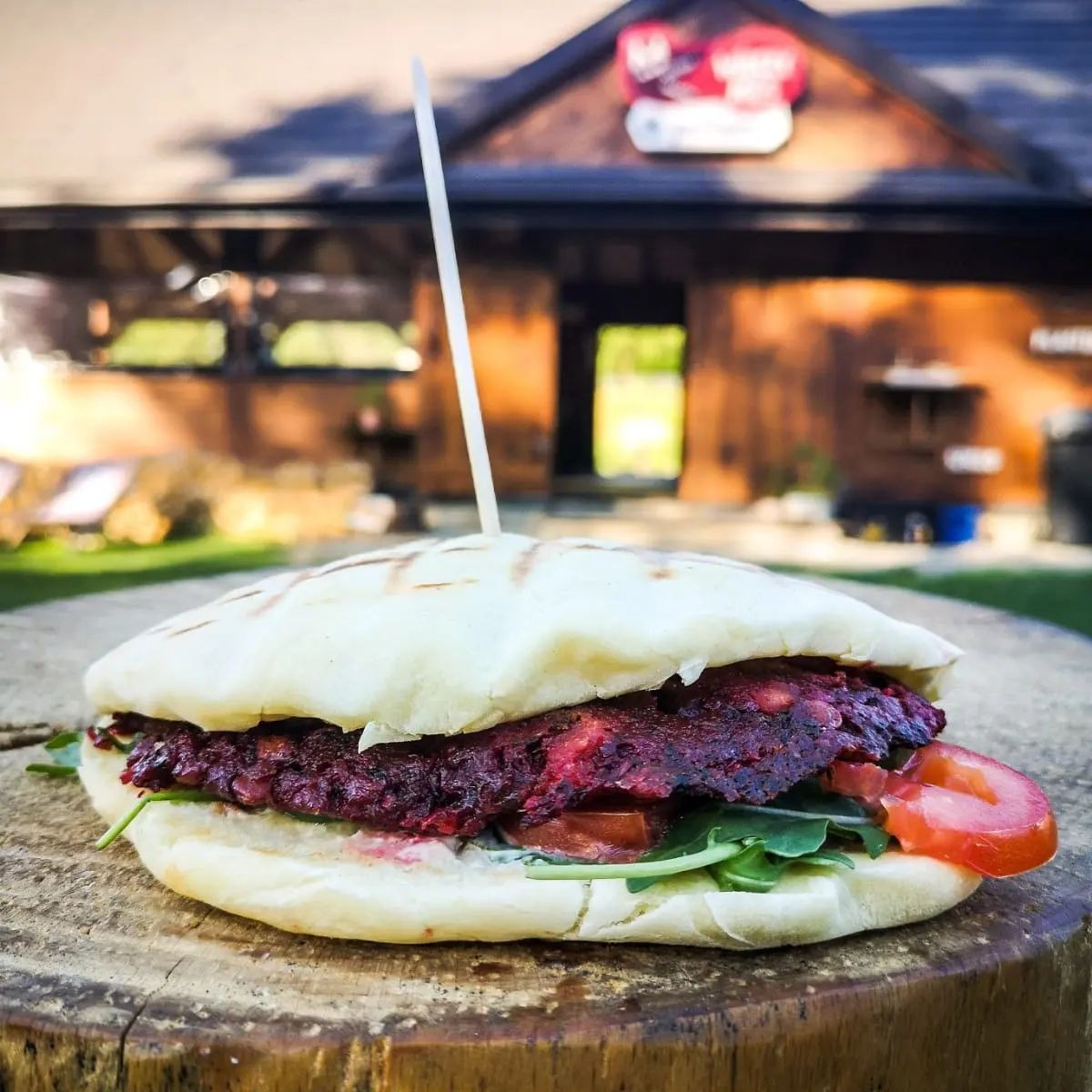 Burger z burakiem i warzywami, podany na drewnianym pniu, z budynkiem w tle.