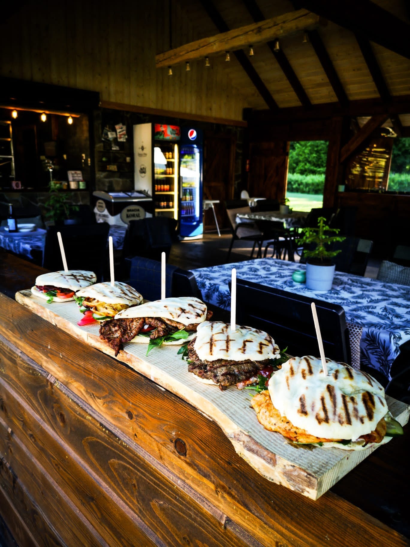 Rząd pięciu kanapek z grilla na drewnianej desce, z widocznymi warstwami mięsa, warzyw i sosów, serwowanych w plenerowej restauracji z drewnianą konstrukcją i stolikami w tle.