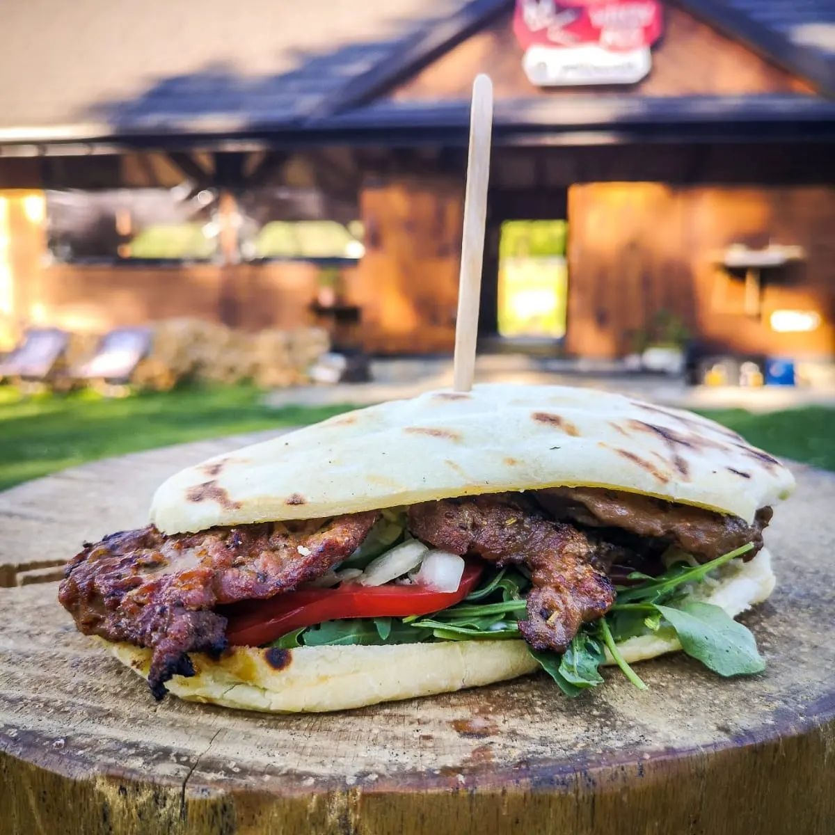 Pita z grillowanym mięsem, rukolą, pomidorem i cebulą, podana na drewnianym pniu, z budynkiem w tle i zielonym trawnikiem.