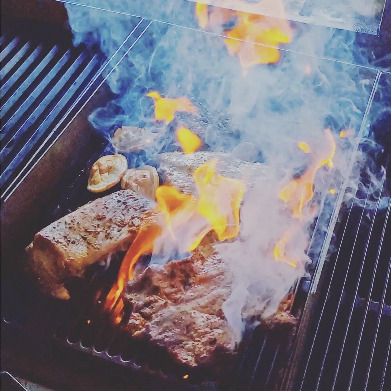 Grillowane mięso i warzywa w płomieniach na ruszcie, widoczne przyprawy i dym, ujęcie z góry.