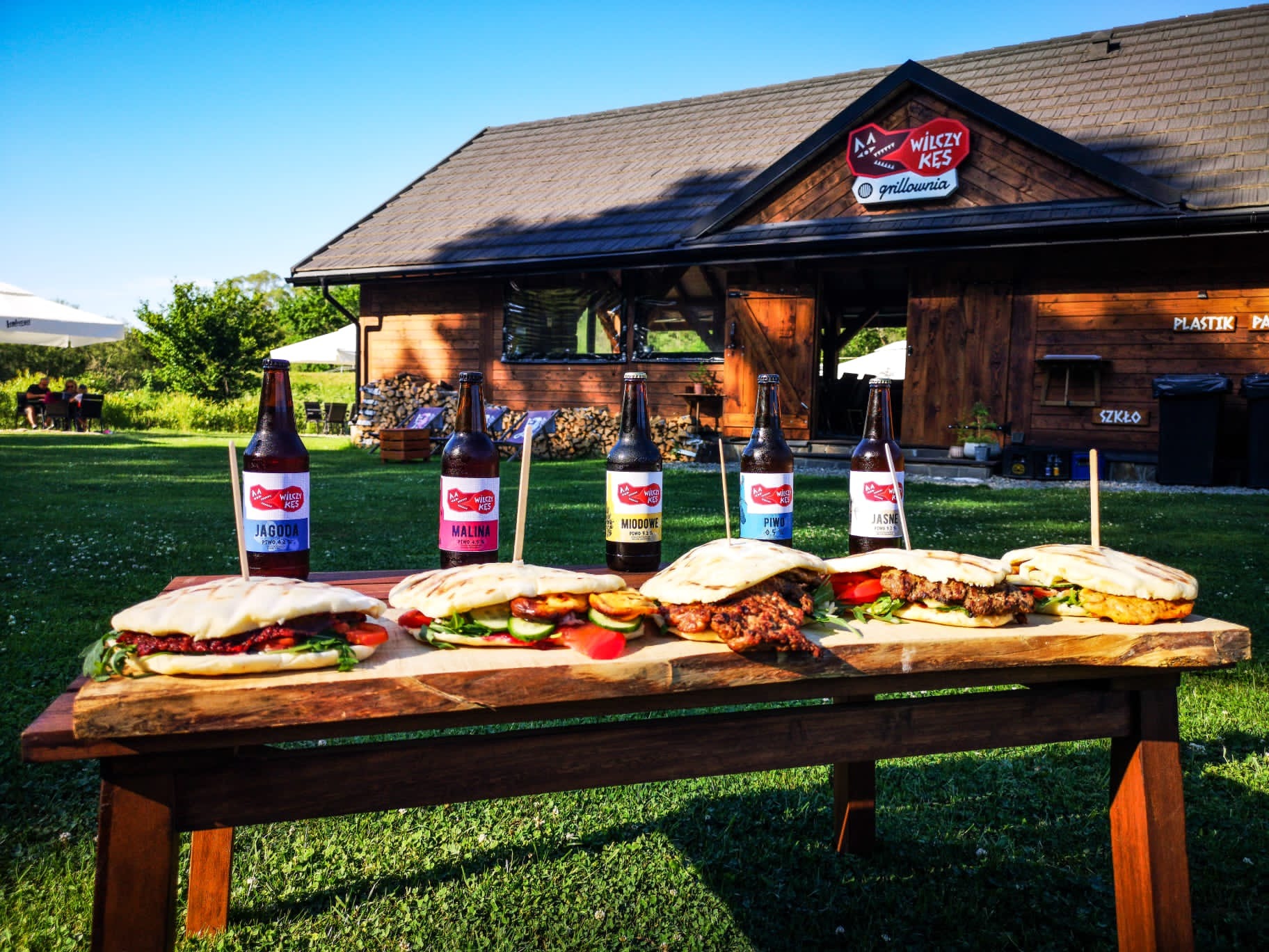 Pięć butelek piwa rzemieślniczego z etykietami Jagoda, Malina, Miodowe, Piwo i Jasne, ustawionych za rzędem kanapek pita na drewnianym stole, na tle drewnianej grillowni Wilczy Kęs i zielonego...
