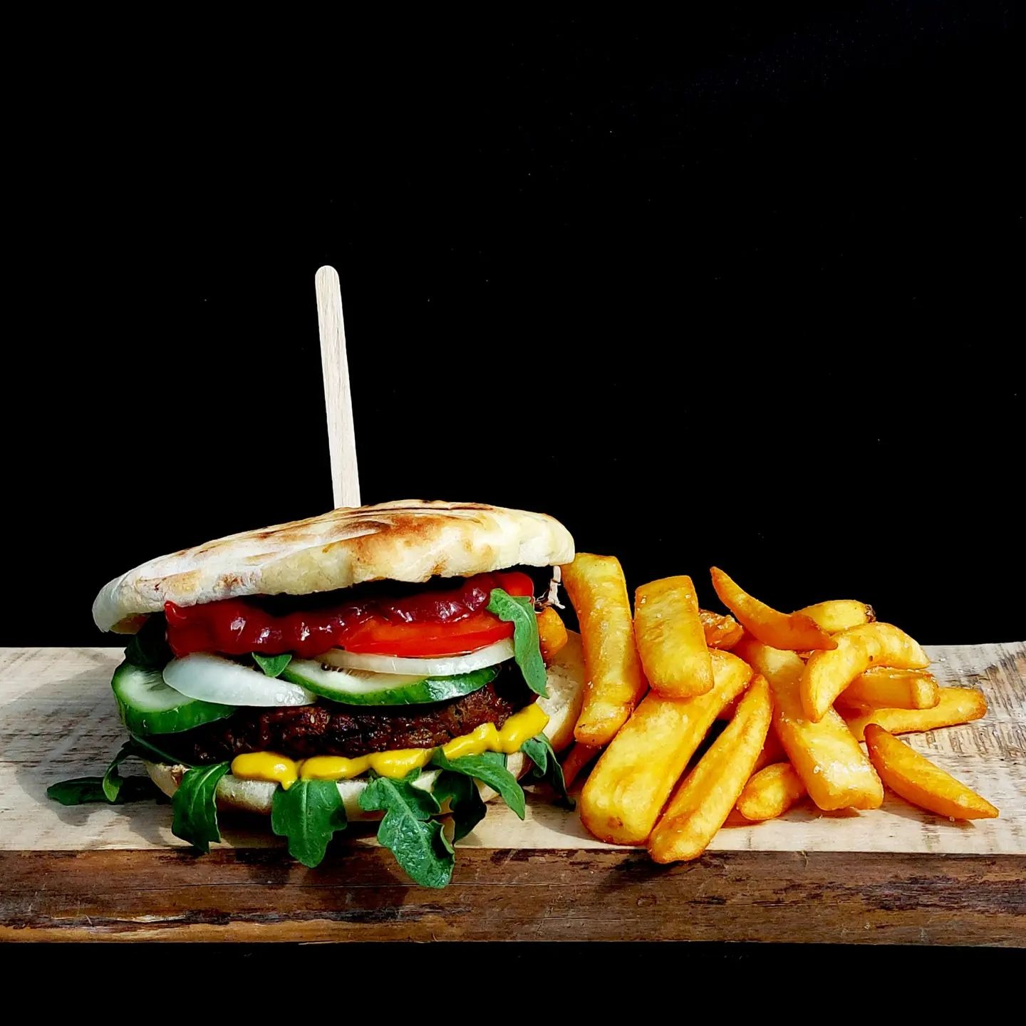 Burger z warzywami, sosem i frytkami na drewnianej desce, na czarnym tle.