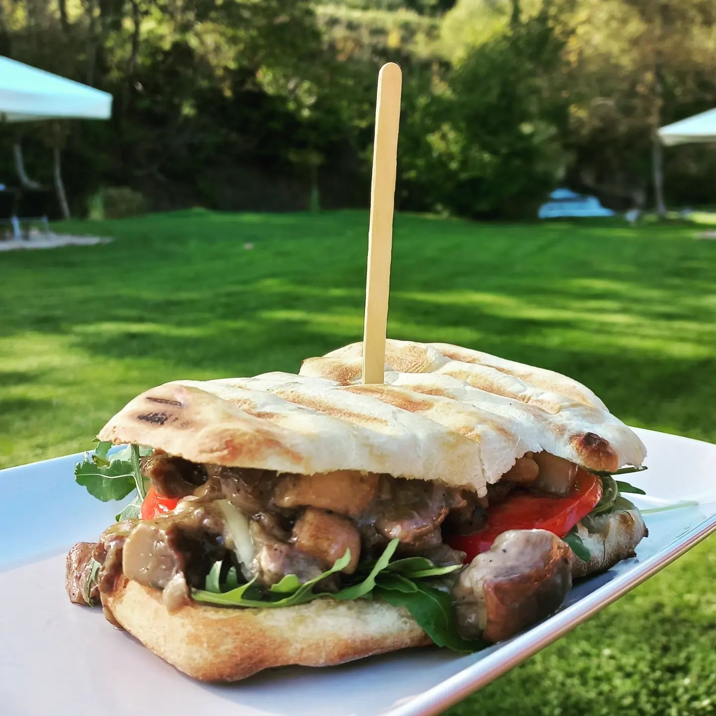 Grillowana kanapka z pieczarkami, pomidorem i rukolą, spięta drewnianym patyczkiem, na białym talerzu, w plenerze na tle zielonego trawnika i drzew.