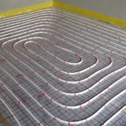 Białe rury ogrzewania podłogowego ułożone na siatce z czerwonymi kwadratami, zabezpieczone żółtą folią przy ścianie.