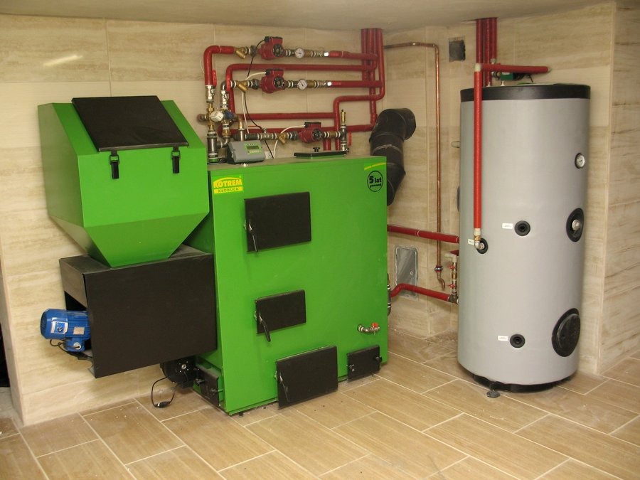 Zielony piec Kotrem z podajnikiem i szary zasobnik ciepłej wody użytkowej, instalacja grzewcza z czerwonymi rurami w kotłowni wyłożonej jasnymi płytkami.