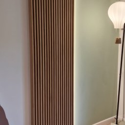 Yurii Leniv - Drewniana ażurowa ścianka działowa z oświetleniem LED, zamontowana na styku białej i pistacjowej ściany, uzupełniona lampą w kształcie balonu na tle parkietu.