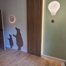 Yurii Leniv - Pokój dziecięcy z drewnianą podłogą, panelami ściennymi z listewek i lampą w kształcie balonu na tle zielonej ściany, uzupełniony dekoracjami ściennymi z motywem niedźwiedzi.