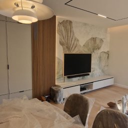 Yurii Leniv - Nowoczesny salon w Warszawie po remoncie: zabudowa meblowa z szarymi frontami, drewniane lamele dekoracyjne obok telewizora, fototapeta z motywem roślinnym. Krzesła zabezpieczone folią ochronną.