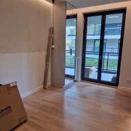 Yurii Leniv - Pokój po remoncie z drewnianą podłogą, częściowo wyszpachlowaną ścianą, lustrem wbudowanym w ścianę i kartonem na podłodze.