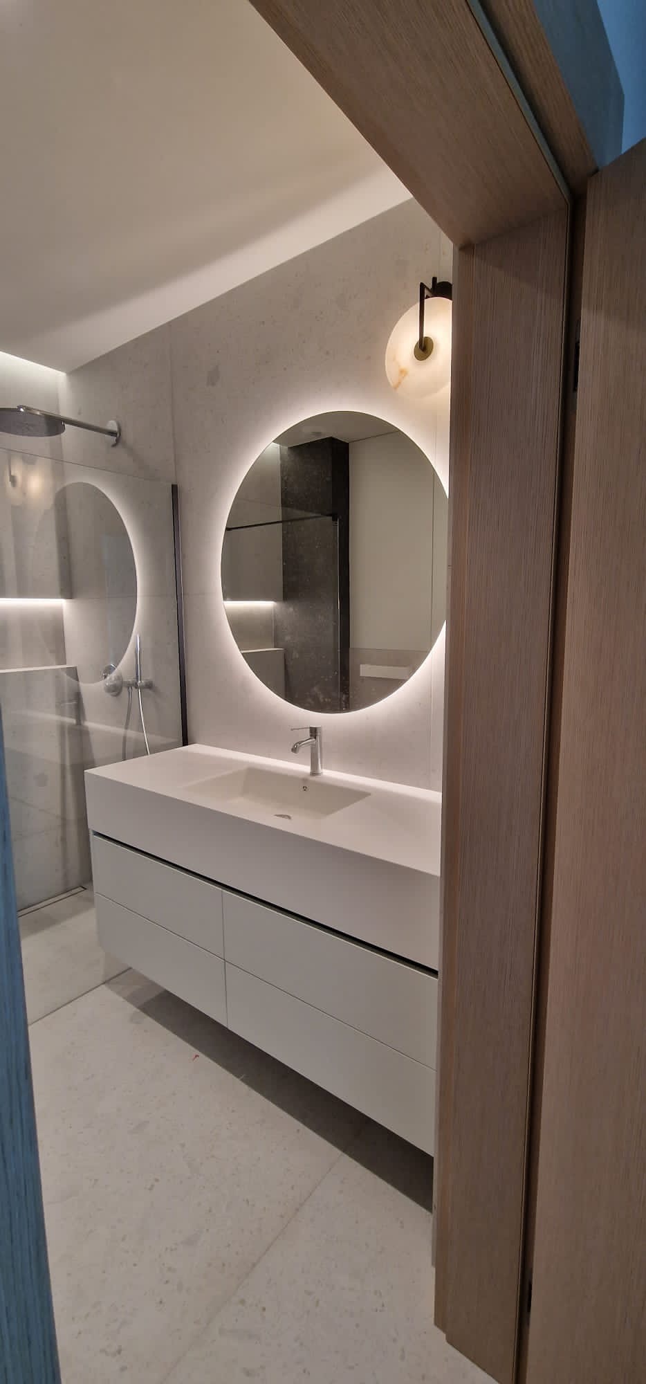 Nowoczesna łazienka z białymi szafkami pod umywalką, okrągłym lustrem z podświetleniem LED i szklaną kabiną prysznicową, widziana zza drewnianej ościeżnicy.