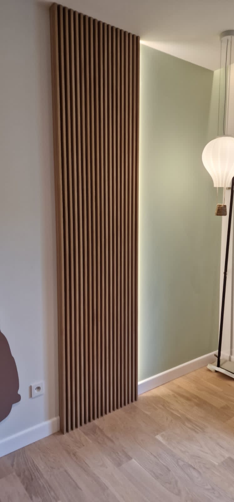 Drewniana ażurowa ścianka działowa z oświetleniem LED, zamontowana na styku białej i pistacjowej ściany, uzupełniona lampą w kształcie balonu na tle parkietu.