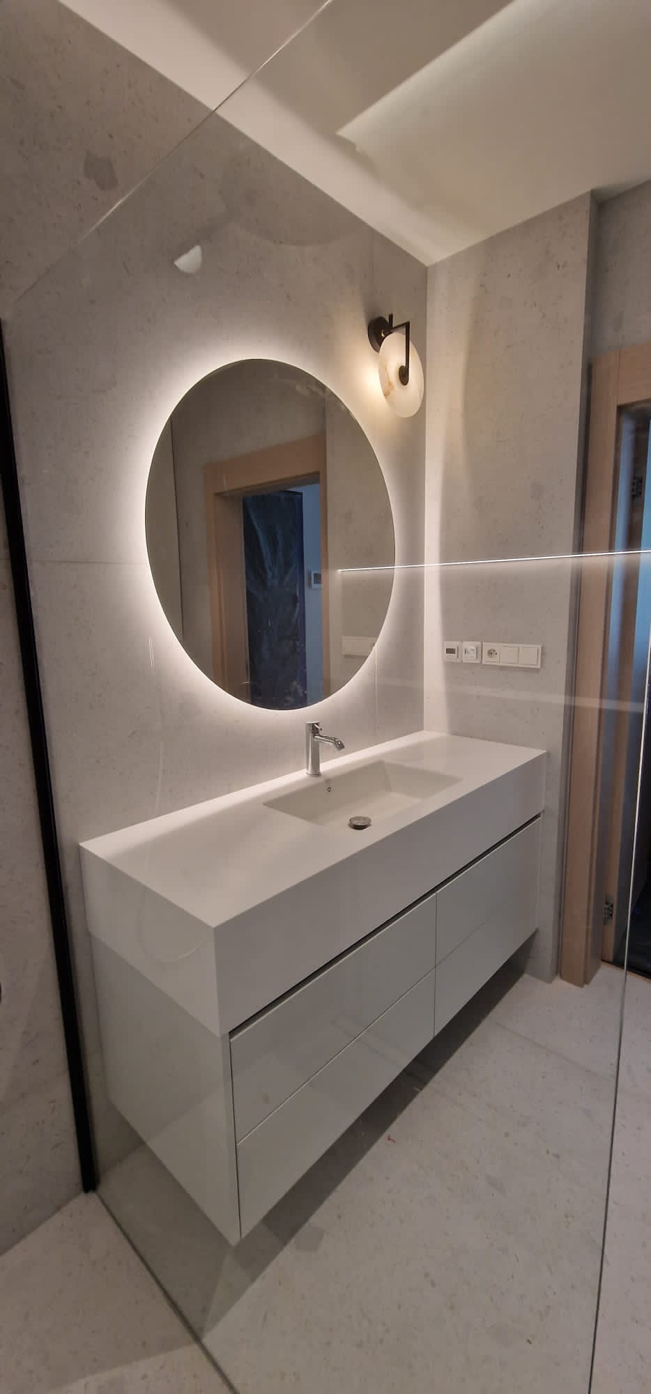 Nowoczesna łazienka z okrągłym lustrem podświetlanym LED, białą szafką pod umywalkę z szufladami i szarą płytką imitującą kamień na ścianach.