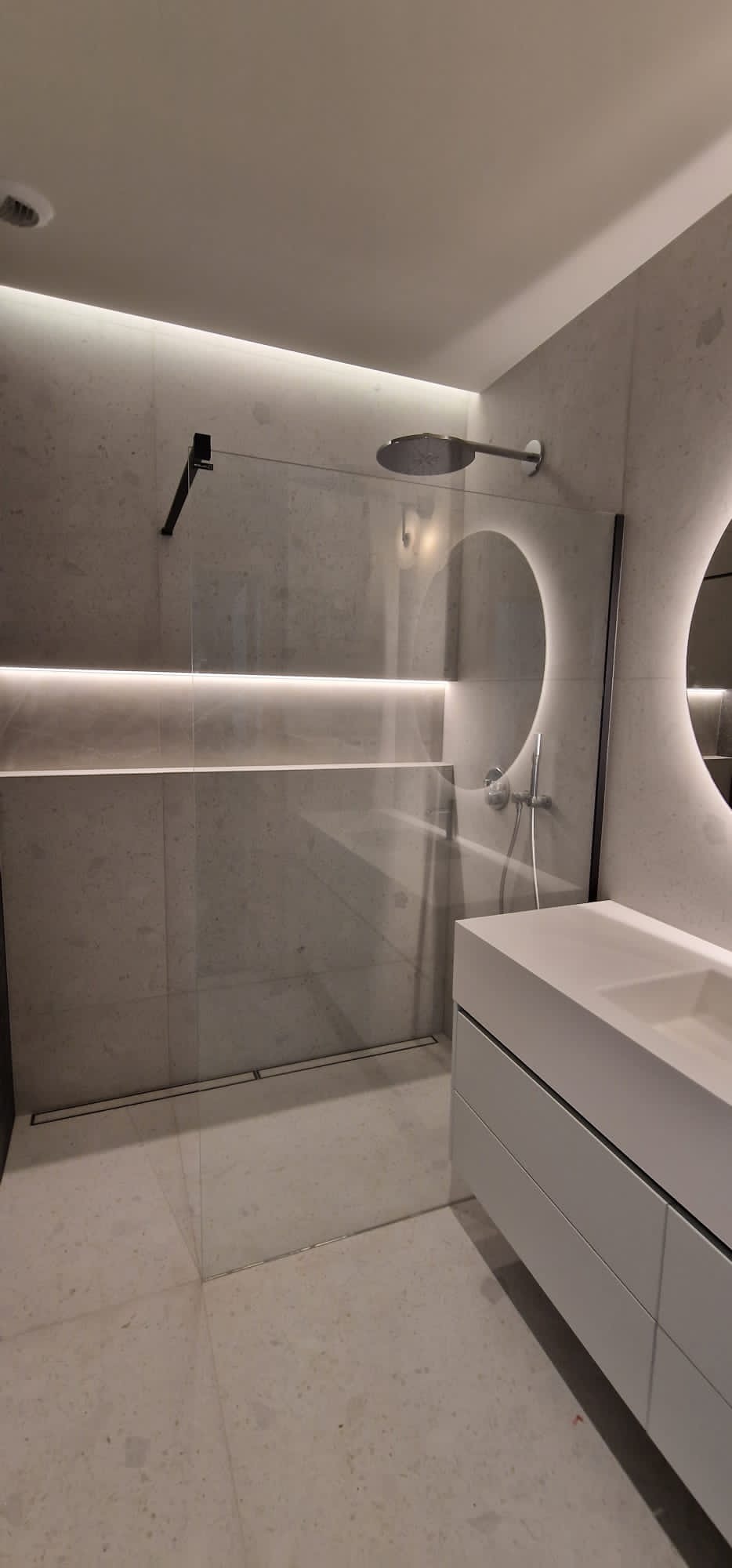 Nowoczesna łazienka z kabiną prysznicową typu walk-in, podświetlaną półką, okrągłym lustrem z podświetleniem LED i białą szafką z umywalką.