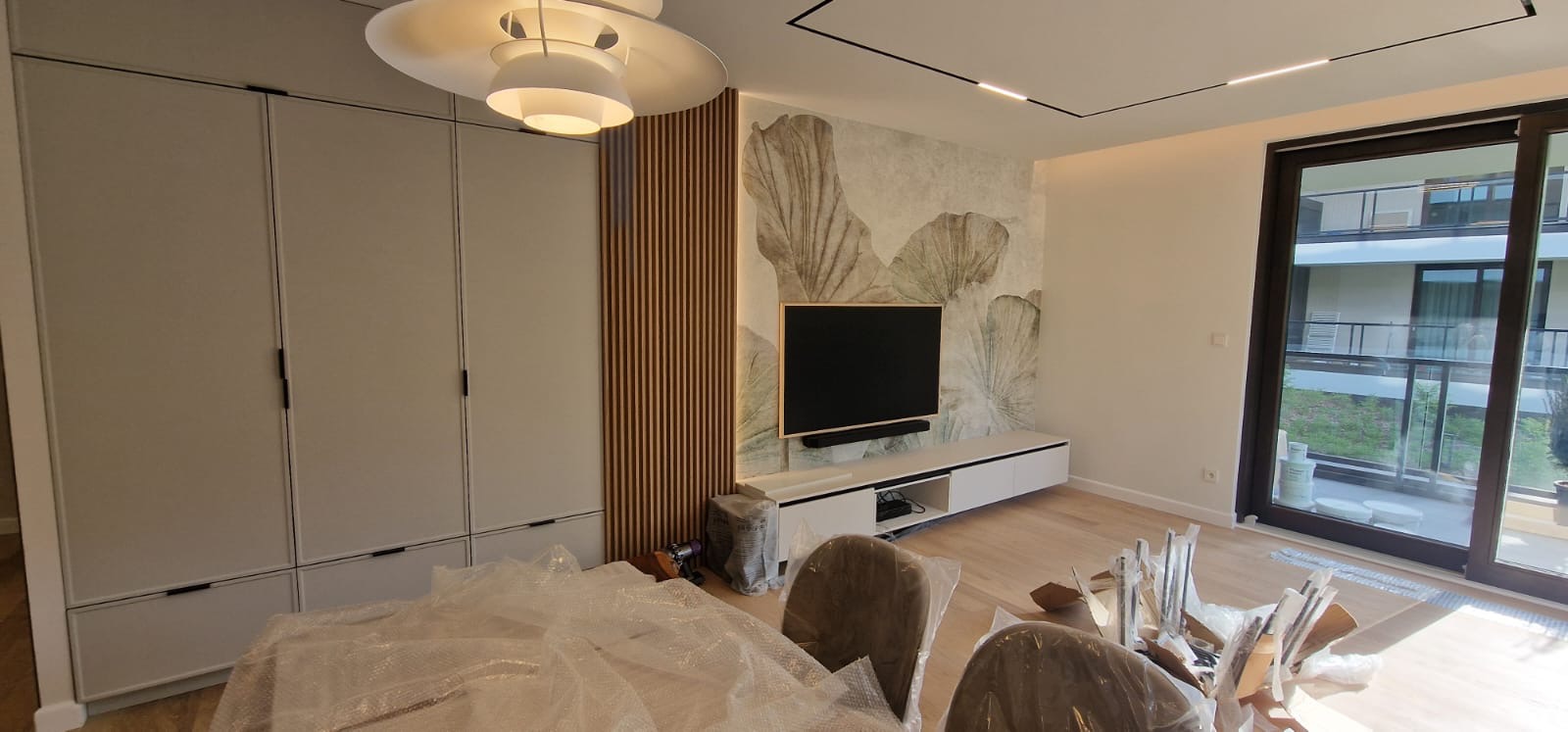 Nowoczesny salon w Warszawie po remoncie: zabudowa meblowa z szarymi frontami, drewniane lamele dekoracyjne obok telewizora, fototapeta z motywem roślinnym. Krzesła zabezpieczone folią ochronną.