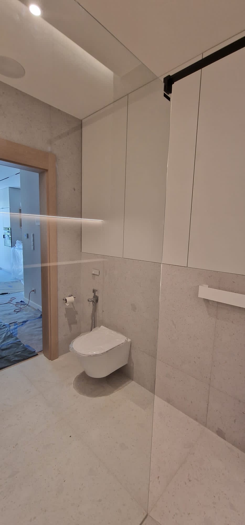 Nowoczesna łazienka z podwieszaną toaletą w trakcie montażu, zabezpieczona folią ochronną, widoczne białe szafki wiszące i szklana kabina prysznicowa.