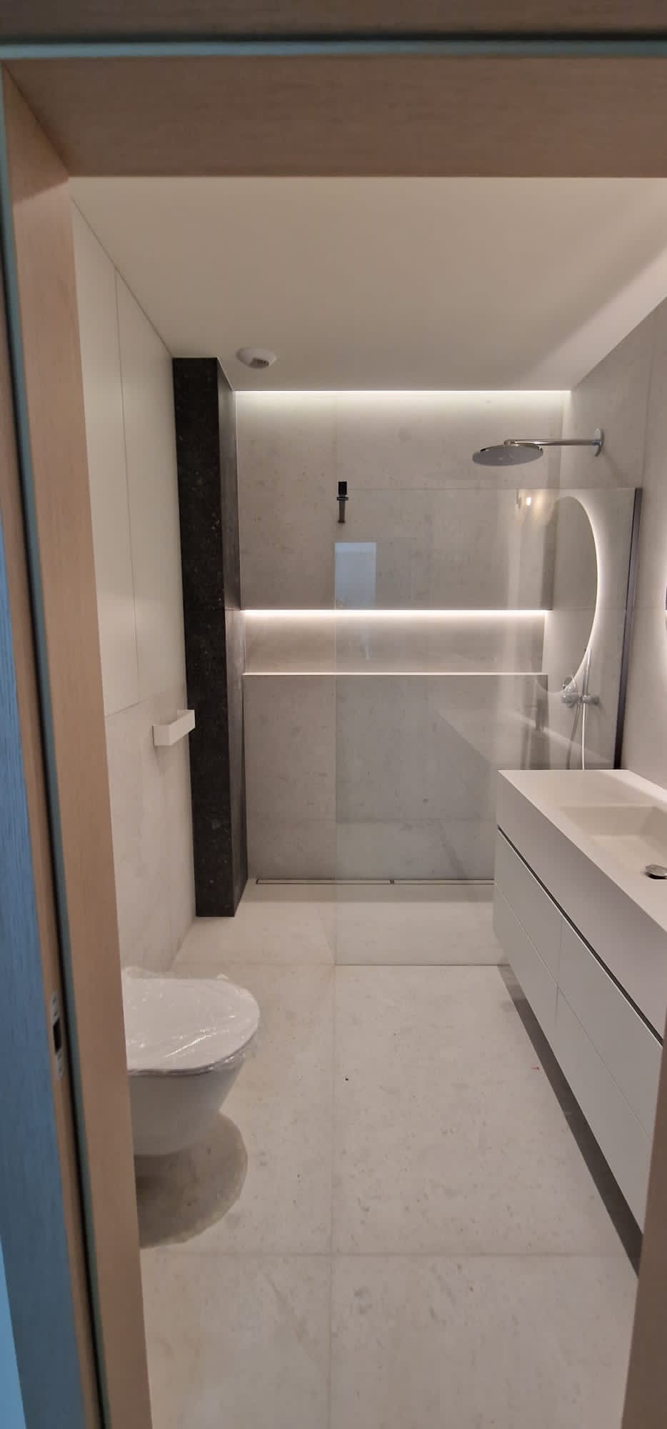 Nowoczesna łazienka z kabiną prysznicową typu walk-in z podświetleniem LED, umywalką nablatową z szafką, i toaletą z folią ochronną na desce, widok zza otwartych drzwi.