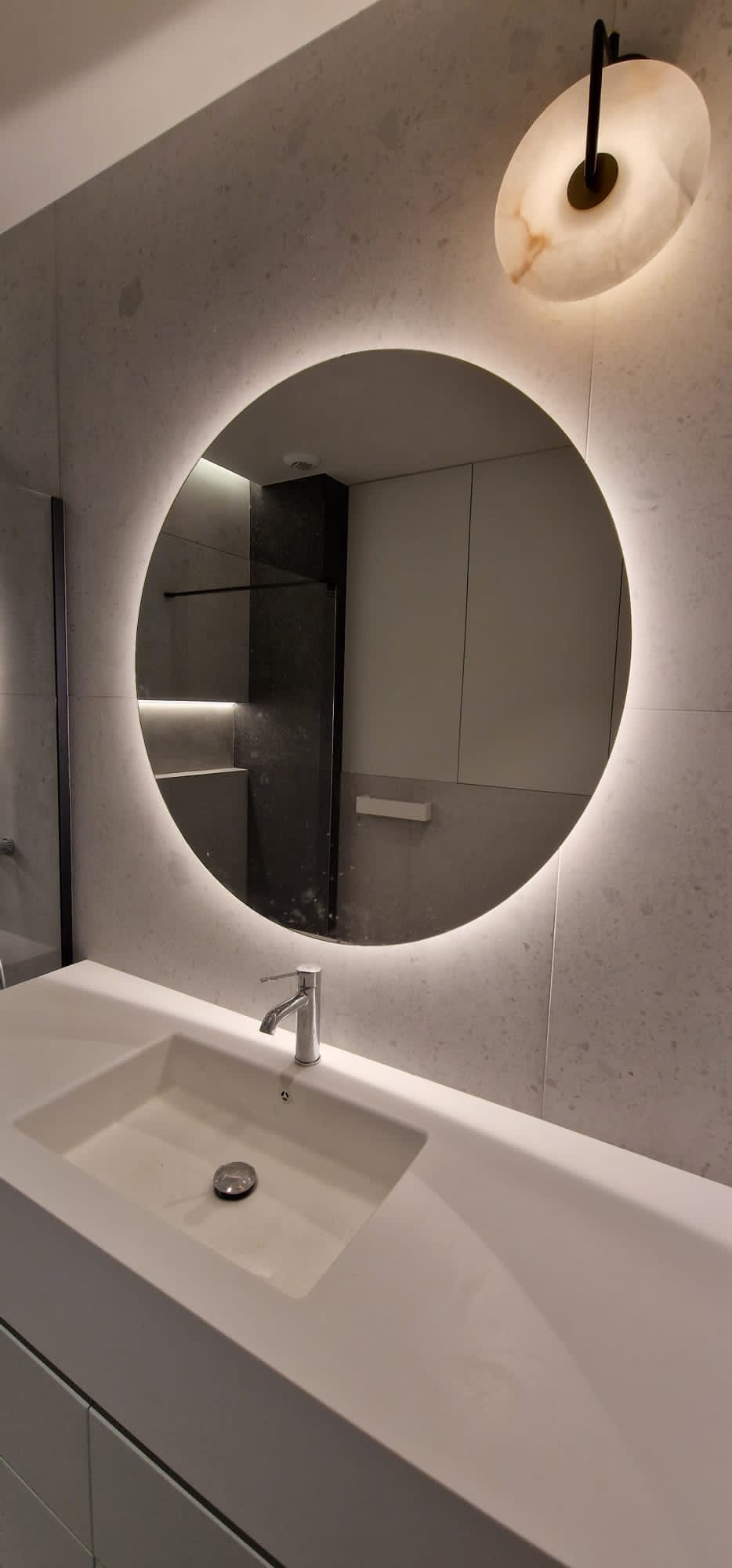 Nowoczesna łazienka z okrągłym lustrem podświetlanym LED, umywalką wpuszczaną w blat i marmurową lampą ścienną.