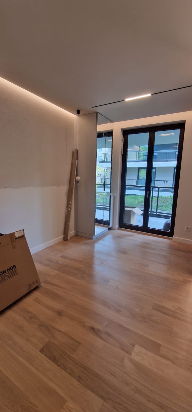 Pokój po remoncie z drewnianą podłogą, częściowo wyszpachlowaną ścianą, lustrem wbudowanym w ścianę i kartonem na podłodze.