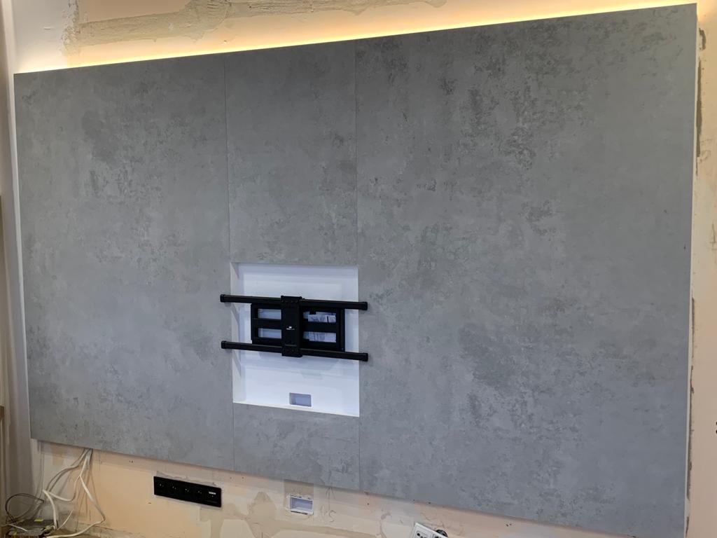Szara ściana z efektem betonu, podświetlona od góry listwą LED, z zamontowanym uchwytem do telewizora w białej wnęce. Widoczne niedokończone prace wykończeniowe u dołu ściany.