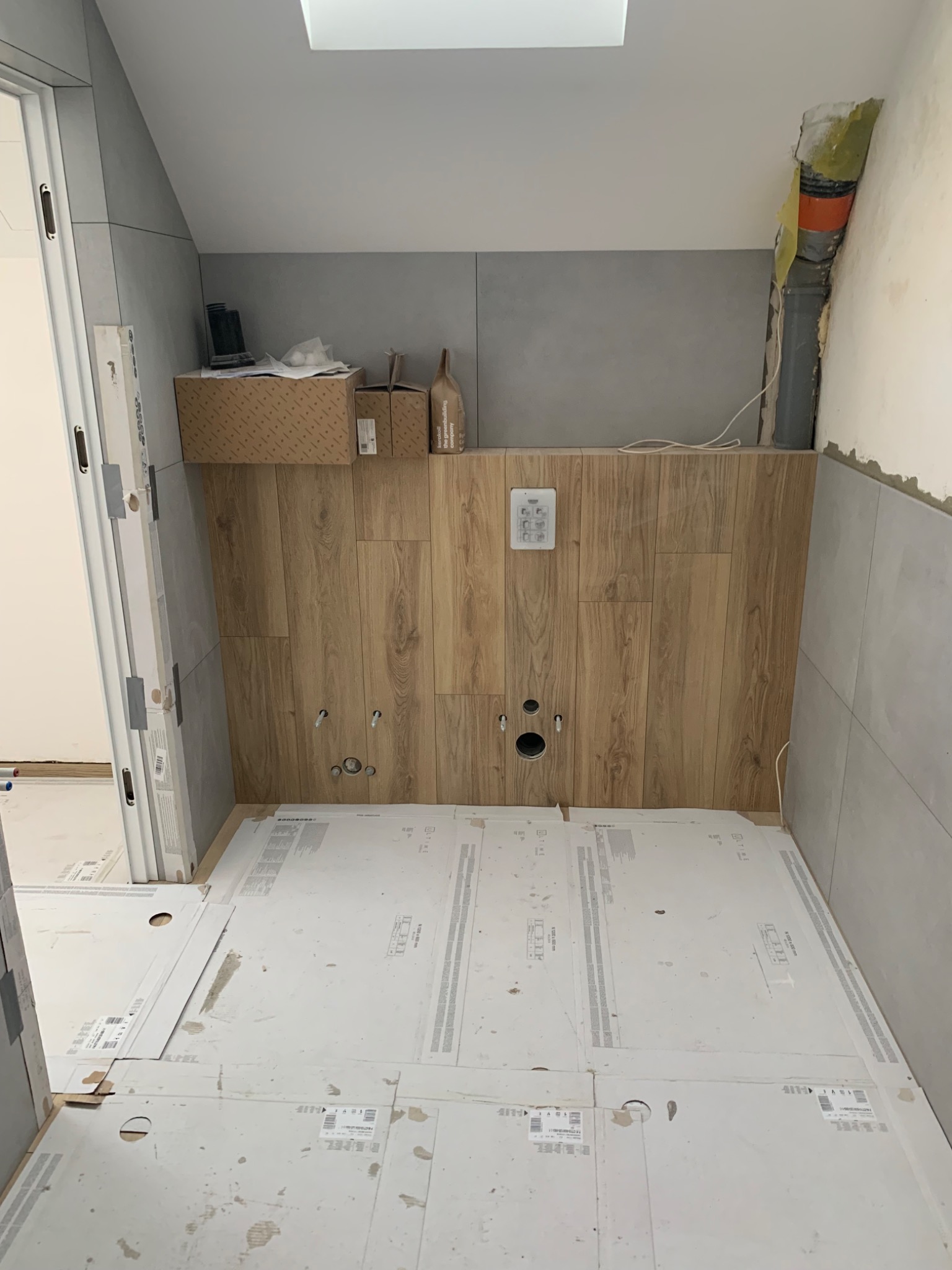 Łazienka w trakcie remontu: ściany wyłożone szarymi płytkami i panelami imitującymi drewno, zabezpieczona podłoga, widoczne otwory na armaturę, instalacja kanalizacyjna w rogu.