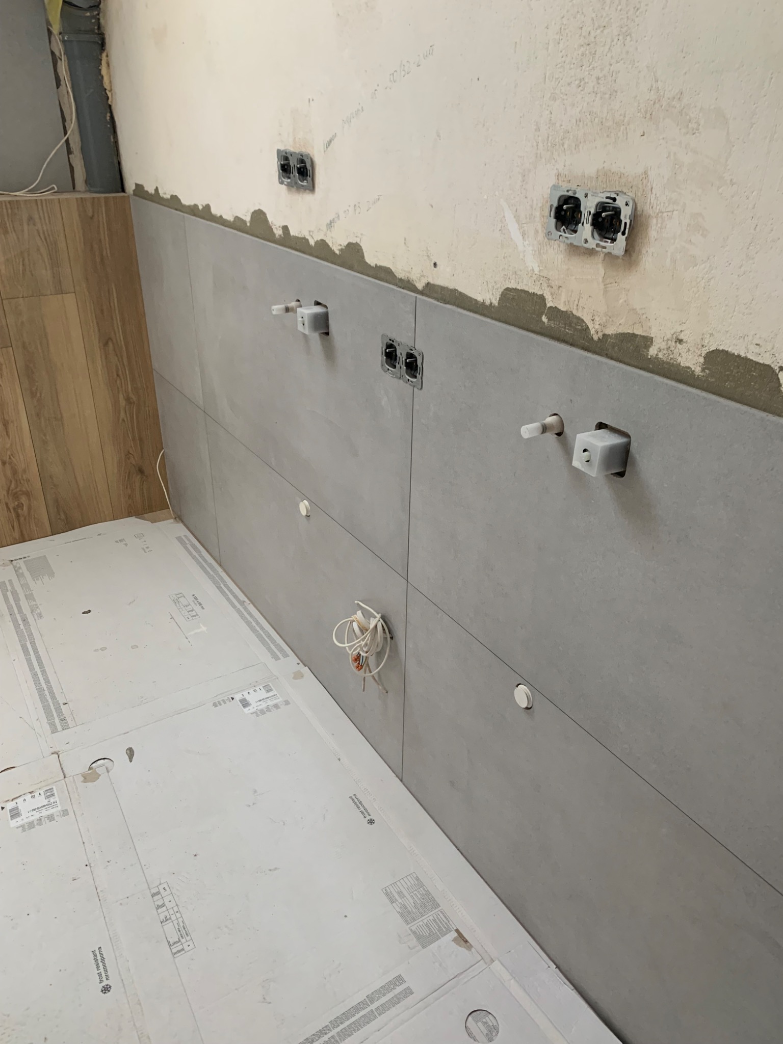 Montaż dużych, szarych płytek ceramicznych na ścianie w łazience, widoczne wystające kable i gniazdka elektryczne, podłoga zabezpieczona kartonem.