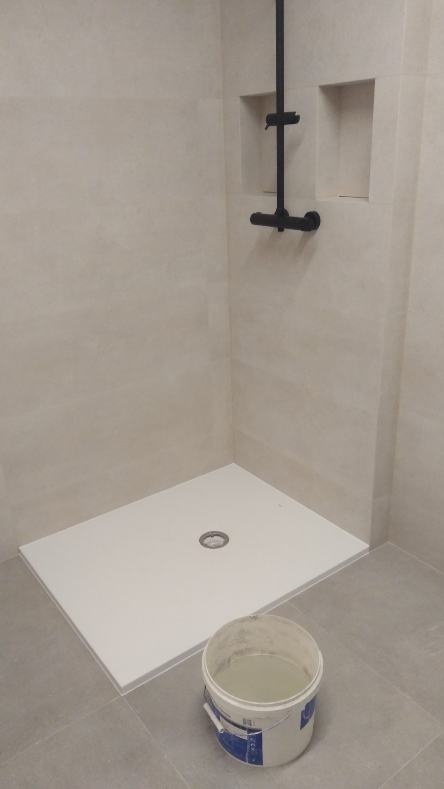 Wykończona łazienka w minimalistycznym stylu: biały brodzik z odpływem w posadzce, czarna armatura prysznicowa, beżowe płytki ścienne z wnęką, wiadro z wodą na szarych płytkach podłogowych.