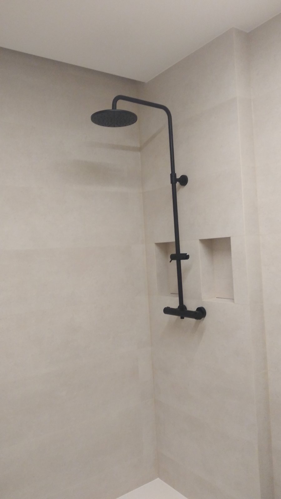 Nowoczesna łazienka z beżowymi płytkami i czarną armaturą: deszczownica i bateria zamontowane na ścianie, w ścianie wnęka.