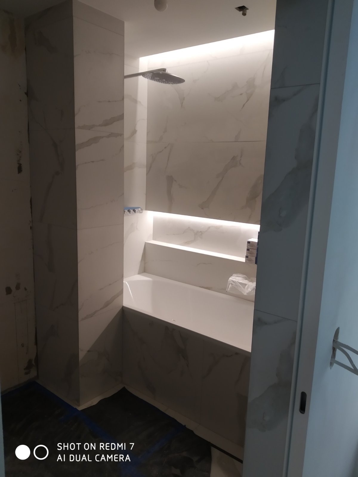 Nowoczesna łazienka w trakcie remontu, widoczna wanna obudowana płytkami imitującymi marmur, podświetlona wnęka z półkami, surowy stan wykończenia podłogi.