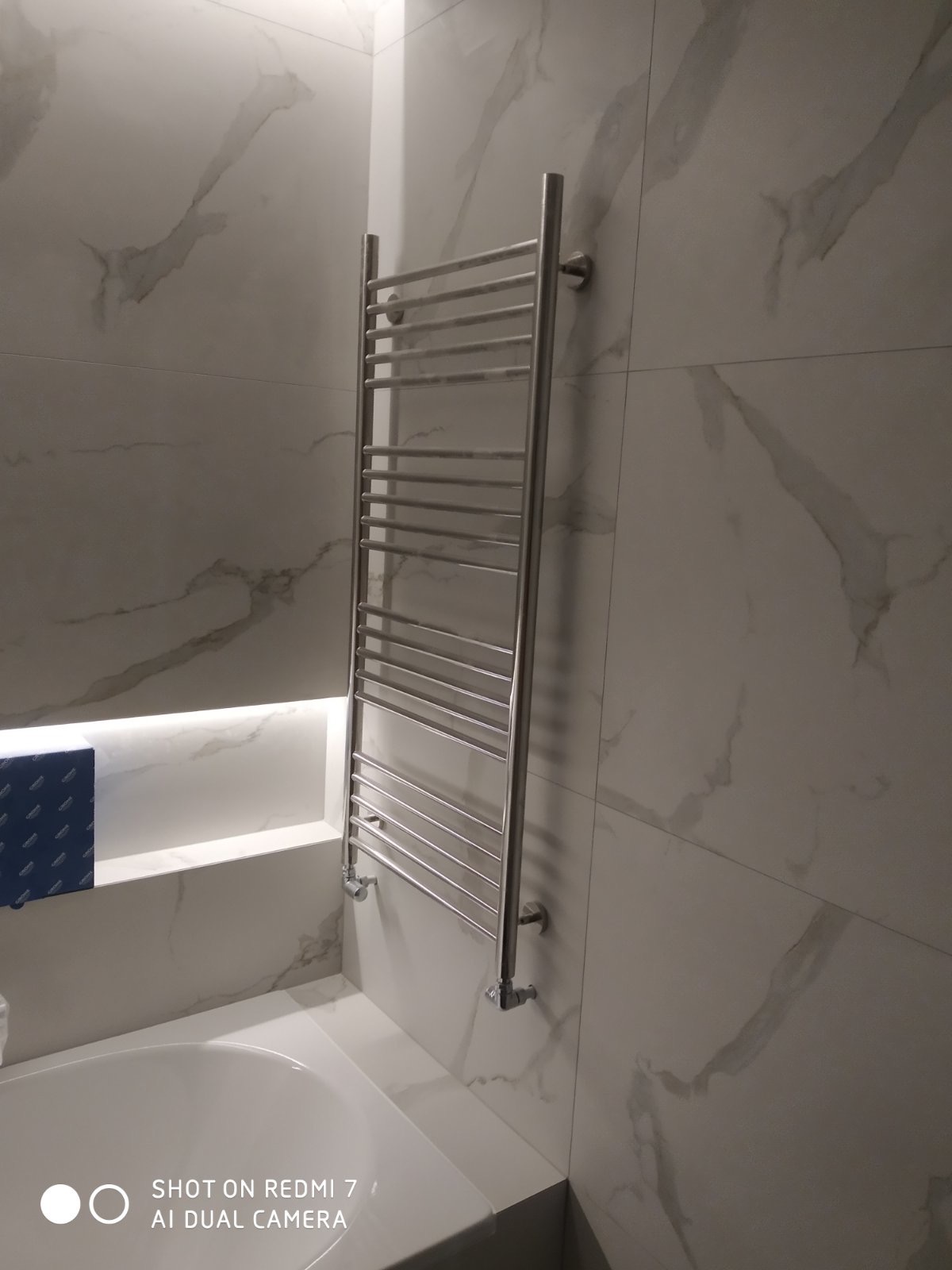 Chromowany grzejnik drabinkowy zamontowany na ścianie z płytek imitujących marmur w łazience, widoczny fragment wanny i podświetlona wnęka.