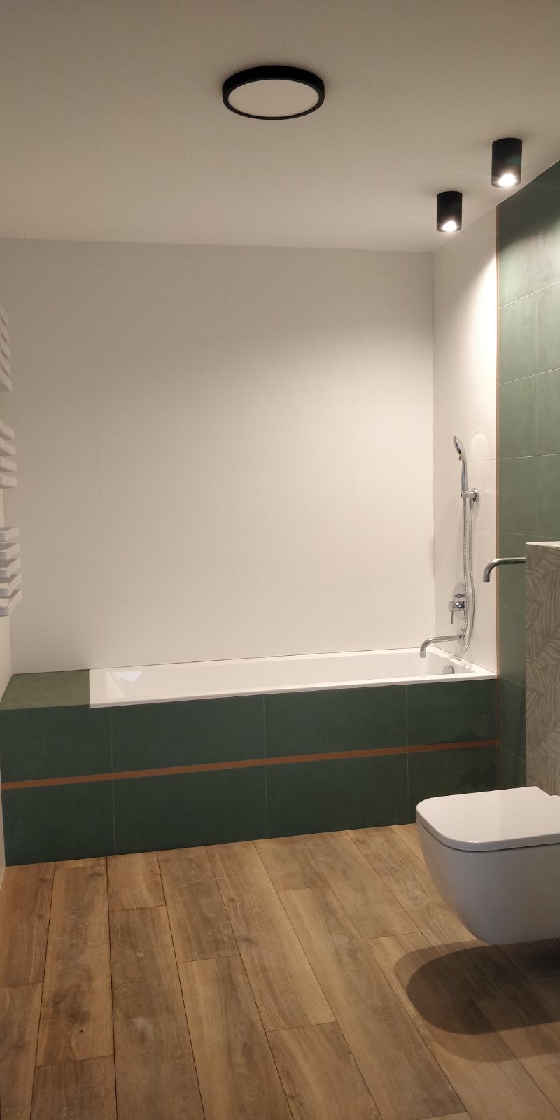 Nowoczesna łazienka z wanną obudowaną zielonymi płytkami z brązowym akcentem, drewnopodobna podłoga, biała ceramika i oświetlenie punktowe.