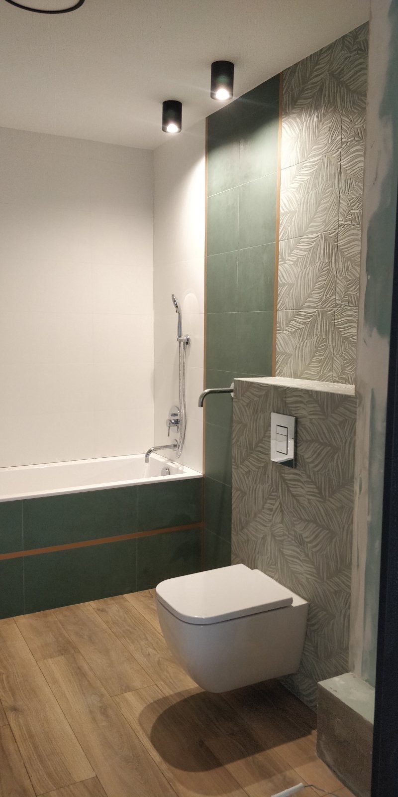 Nowoczesna łazienka z podwieszaną toaletą, zielonymi i wzorzystymi płytkami oraz drewnopodobną podłogą, oświetlona dwoma punktowymi lampami sufitowymi, widok na wannę i baterię.