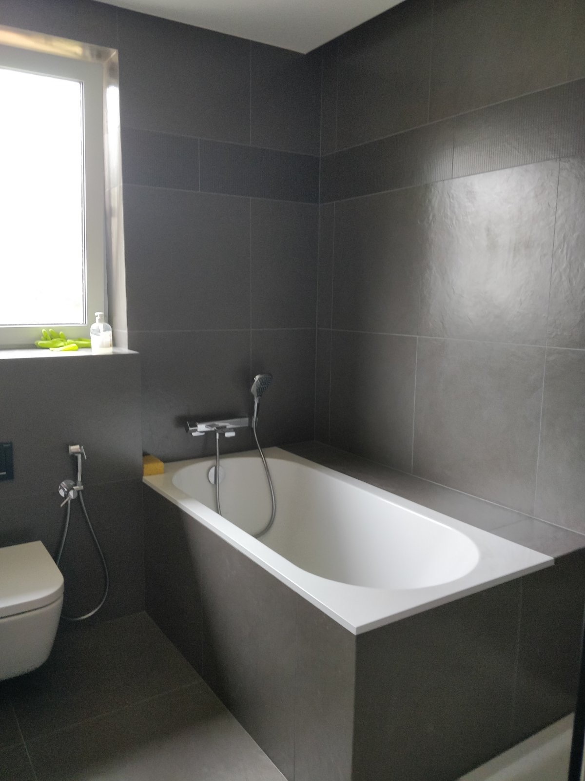 Nowoczesna łazienka z wanną w zabudowie, ciemnoszare płytki na ścianach i podłodze, biała ceramika, okno z zielonymi rękawicami i butelką płynu do dezynfekcji na parapecie.