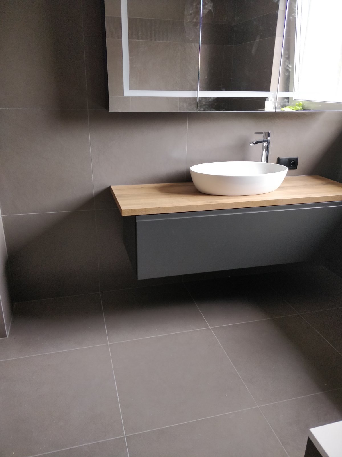 Nowoczesna łazienka z szarą glazurą, białą umywalką nablatową na drewnianym blacie i grafitową szafką pod umywalką, oświetlonym lustrem z szafką oraz gniazdkiem elektrycznym obok baterii.