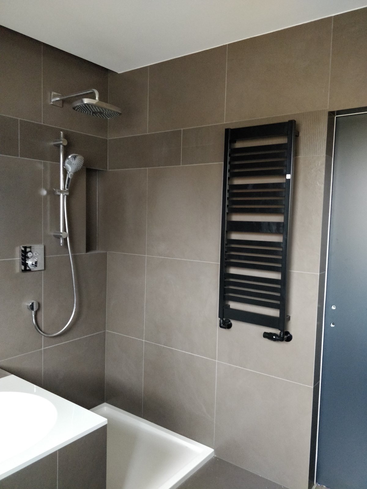 Nowoczesna łazienka z beżowymi płytkami, czarnym grzejnikiem drabinkowym, białą wanną i umywalką, oraz chromowaną armaturą prysznicową.