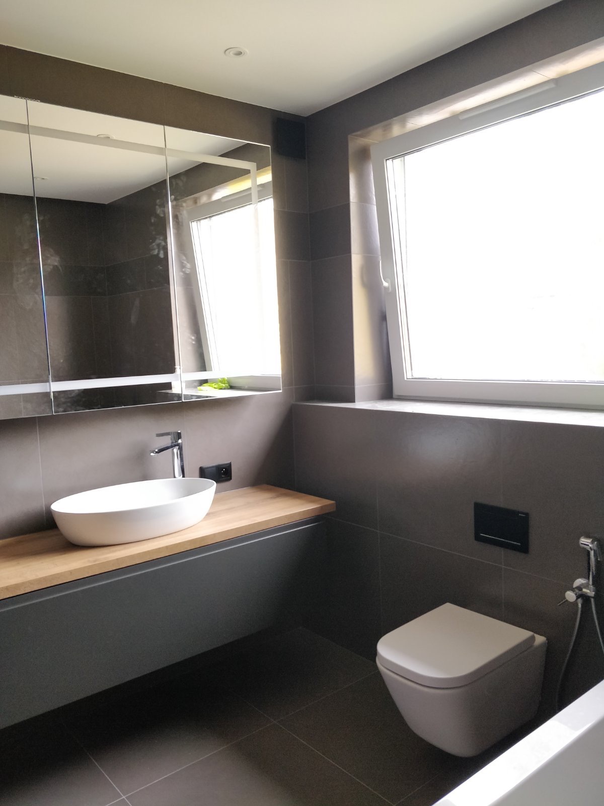 Nowoczesna łazienka w odcieniach szarości z drewnianym blatem pod umywalką, wisząca toaleta i lustro z szafką, oświetlona naturalnym światłem wpadającym przez okno.